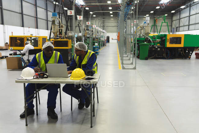 Front view of a middle aged mixed race male factory worker wearing glasses and a Yong African American male factory worker sitting at a table talking and using a laptop computer in a warehouse at a factory processing plant — Stock Photo