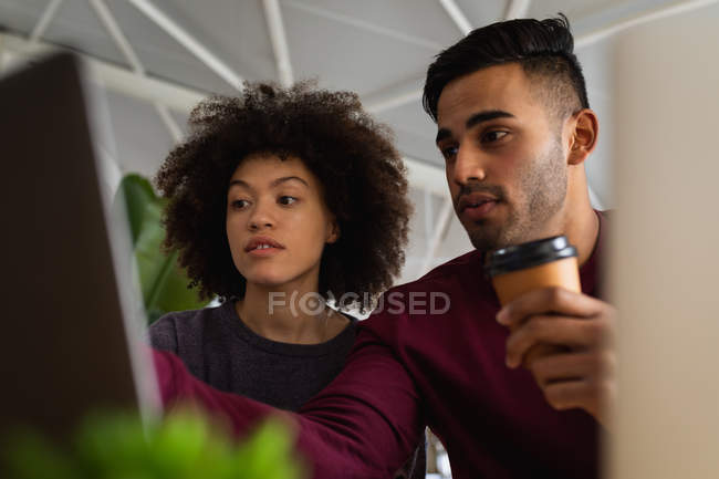 Vue de face gros plan d'un jeune homme de race mixte et d'une jeune femme de race mixte assis à un bureau à l'aide d'ordinateurs portables et regardant un écran dans un bureau créatif — Photo de stock