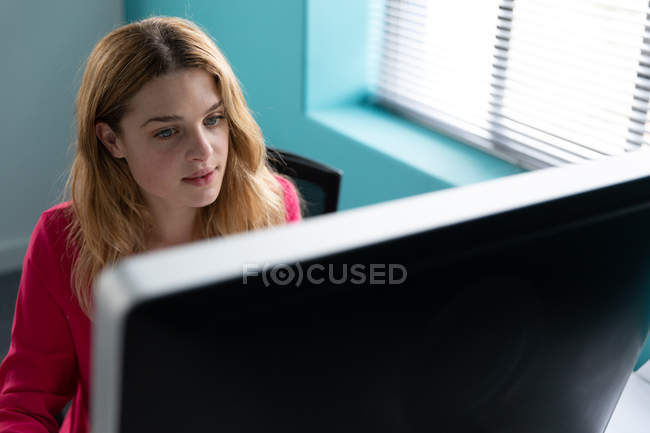 Vista frontal de cerca una joven mujer caucásica sentada en un escritorio junto a una ventana usando una computadora en la oficina moderna de un negocio creativo - foto de stock