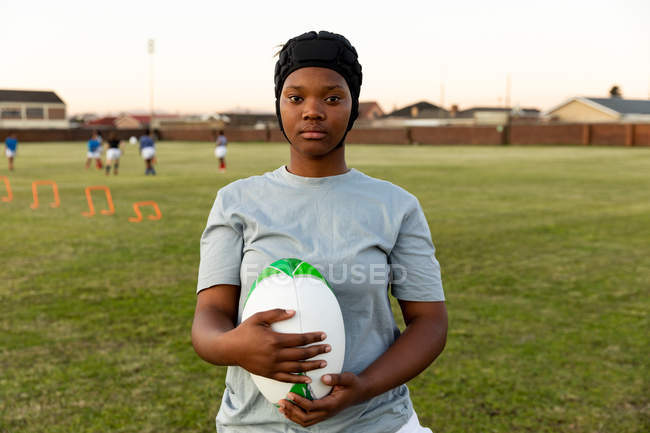 Portrait of a young adult mixed race female rugby player wearing a headguard standing on a sports field holding a rugby ball and looking to camera, with her teammates in the background — Stock Photo