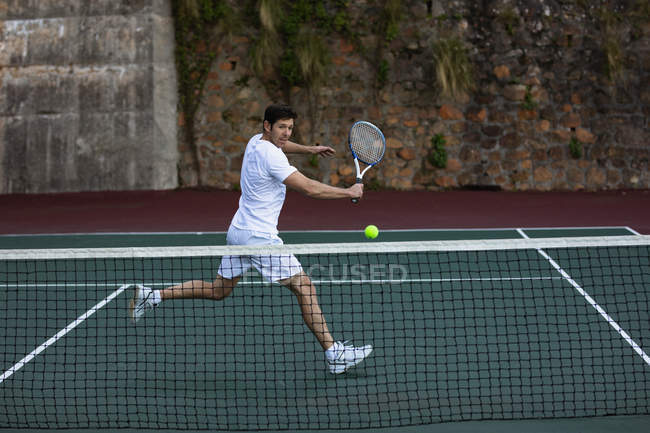 Vista laterale di un giovane caucasico che gioca a tennis, corre verso una palla con un muro alle spalle — Foto stock