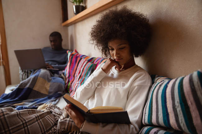 Front view close up of a young mixed race woman sitting on a sofa reading a book at home, her partner, a young African American man, is sitting on the sofa using a laptop computer in the background. — Stock Photo