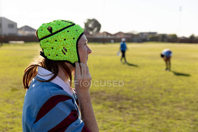 Side view of a young adult Caucasian female rugby player standing on a rugby pitch fastening her headguard, with her teammates in the background — Stock Photo