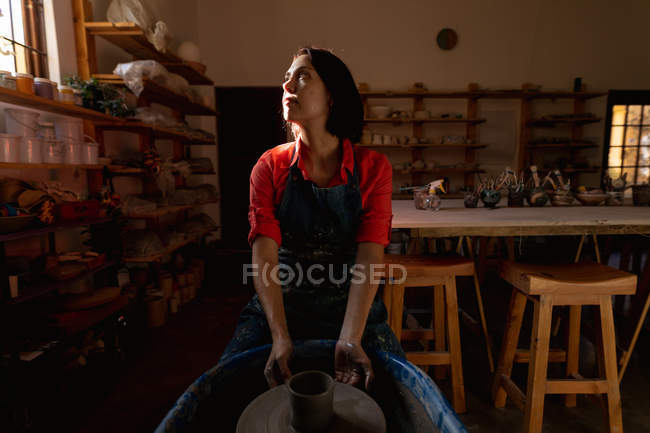 Vista frontal de una joven alfarera caucásica sentada con una olla en una rueda de alfarero en un estudio de cerámica, y mirando hacia la ventana - foto de stock