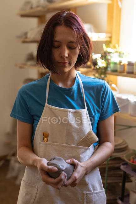 Front view close up of a young Caucasian female potter wearing an apron looking down at a piece of clay she is holding in her hands and modelling in a pottery studio — Stock Photo