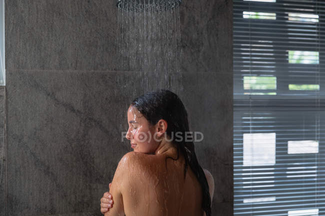 Vue arrière d'une jeune femme brune caucasienne debout sous la douche se lavant les cheveux, la tête tournée vers le côté dans une salle de bain moderne — Photo de stock
