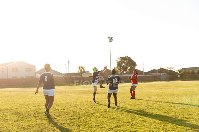 Visão traseira de um grupo de jovens jogadores de rugby femininos adultos multi-étnicos, com a bola no ar durante uma partida de rugby — Fotografia de Stock