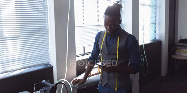 Frontansicht eines jungen afrikanisch-amerikanischen männlichen Modestudenten mit einem Maßband um den Hals, der ein Smartphone in einem Studio der Modehochschule benutzt — Stockfoto