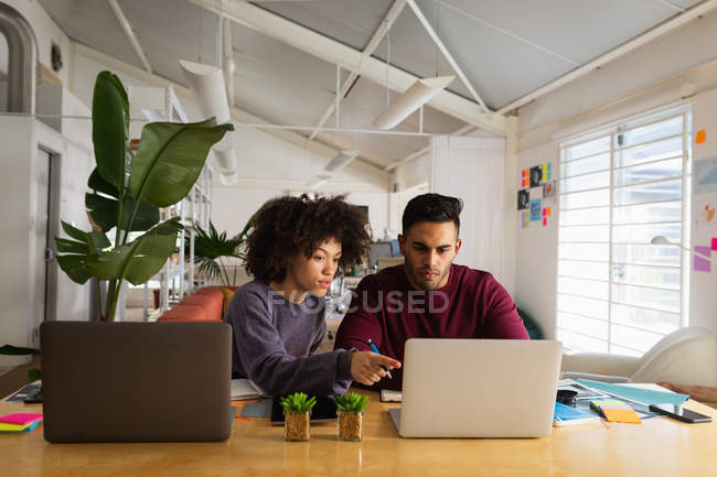 Вид спереду молодого змішаного чоловіка і молодої змішаної раси жінки, що сидить за столом, використовуючи ноутбук комп'ютерів у творчому офісі — стокове фото