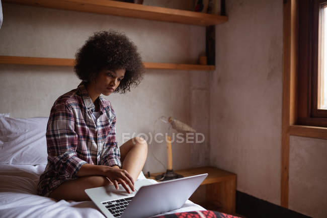 Вид сбоку на молодую женщину смешанной расы, сидящую дома на кровати с ноутбуком — стоковое фото