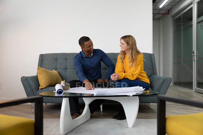 Frontansicht eines jungen afrikanisch-amerikanischen Mannes und einer jungen kaukasischen Frau, die auf einem Sofa im Loungebereich eines modernen Kreativunternehmens miteinander reden und Pläne betrachten — Stockfoto