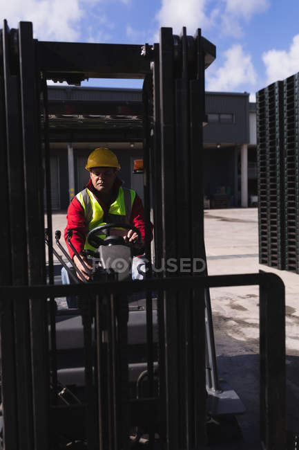 Vue de face gros plan d'un ouvrier d'usine caucasien d'âge moyen utilisant un chariot élévateur pour déplacer une pile de palettes à l'extérieur d'un entrepôt dans une usine — Photo de stock