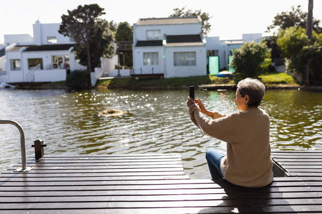 Vista lateral de cerca de una mujer caucásica mayor sentada junto a un lago y tomando fotos con un teléfono inteligente - foto de stock