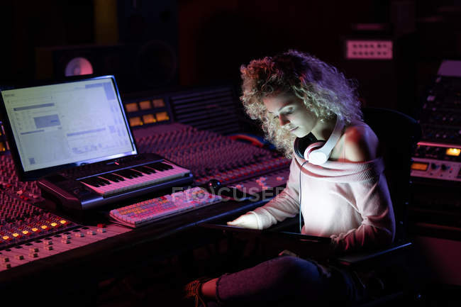 Vista lateral de perto de uma jovem engenheira de som branca sentada e trabalhando em uma mesa de mistura usando um computador portátil em um estúdio de gravação — Fotografia de Stock