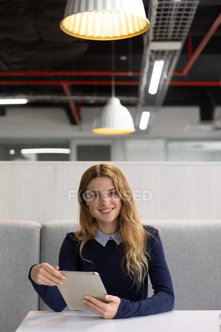 Ritratto ravvicinato di una giovane donna caucasica seduta su una panchina con in mano un tablet a un tavolo nella zona pranzo di un'attività creativa e sorridente alla macchina fotografica — Foto stock