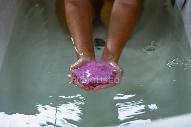 Acercamiento de las manos ventosas de una mujer en un baño con sales de baño rosadas efervescentes en el agua del baño - foto de stock