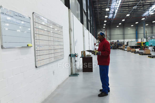 Вид сбоку на афроамериканского работника фабрики среднего возраста, держащего планшет, проверяющий информацию на доске на складе на перерабатывающем заводе — стоковое фото