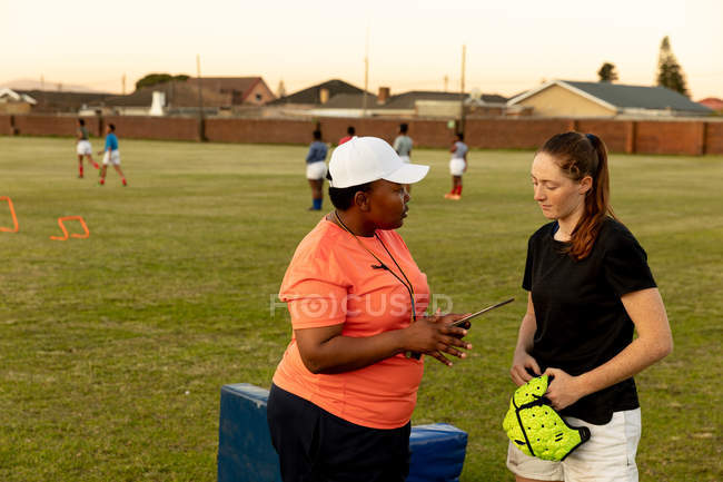 Side view of a middle aged mixed race female rugby coach talking to a young adult Caucasian female rugby player at a sports field during a training session — Stock Photo