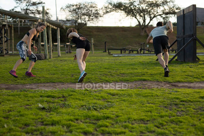 Visão traseira de duas jovens mulheres caucasianas e um jovem caucasiano correndo em um ginásio ao ar livre durante uma sessão de treinamento de bootcamp — Fotografia de Stock