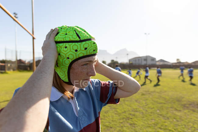 Seitenansicht einer jungen erwachsenen kaukasischen Rugbyspielerin, die auf einem Rugbyfeld steht und ihren Kopfschutz befestigt, während ihre Teamkolleginnen im Hintergrund stehen — Stockfoto