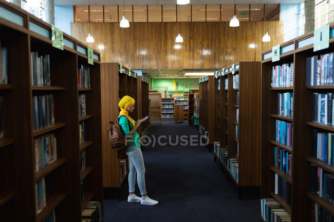 Бічний вид на молоду азіатську студентку, яка носить хіджаб, використовуючи смартфон у бібліотеці. — стокове фото
