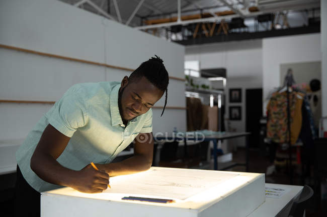 Вид сбоку на молодого афроамериканского студента-модельера, работающего над дизайнерским рисунком на световом ящике в студии колледжа моды — стоковое фото
