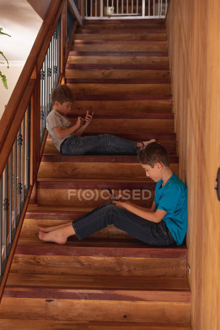 Seitenansicht von zwei vorjugendlichen kaukasischen Jungen, die zu Hause auf einer Treppe sitzen und Smartphones benutzen — Stockfoto