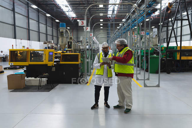 Front view of a middle aged Caucasian female and male factory worker standing in discussion looking at tablet computer between rows of equipment in a warehouse at a processing plant — Stock Photo