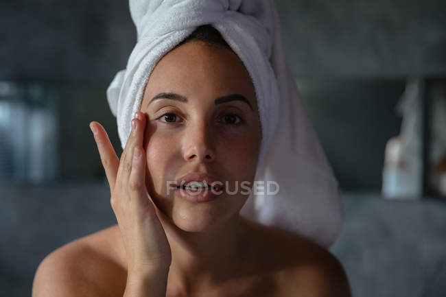 Retrato de cerca de una joven morena caucásica con el pelo envuelto en una toalla, mirando directamente a la cámara y tocándose la cara con una mano en un baño moderno - foto de stock