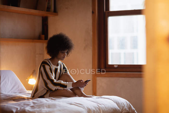 Vista lateral de uma jovem mulher de raça mista usando um smartphone sentado em sua cama em casa com a lâmpada de cabeceira ligada, vista da porta — Fotografia de Stock