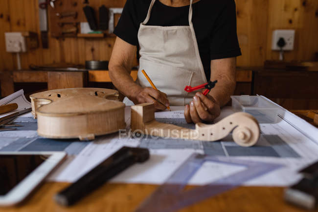 Nahaufnahme einer Geigenbauerin, die in ihrer Werkstatt Notizen macht, mit einer Rolle einer Geige und einem Tablet-Computer vor sich — Stockfoto