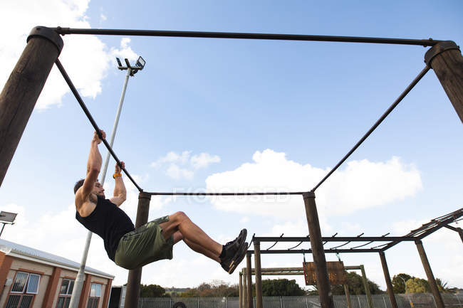 Vista lateral de un joven caucásico balanceándose en un bar en un gimnasio al aire libre durante una sesión de entrenamiento de bootcamp - foto de stock