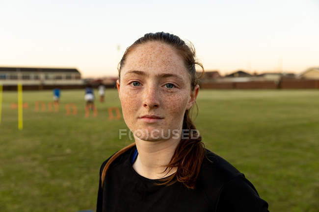Portrait close up of a young adult Caucasian female rugby player standing on a rugby pitch looking to camera, with her teammates training in the background — Stock Photo