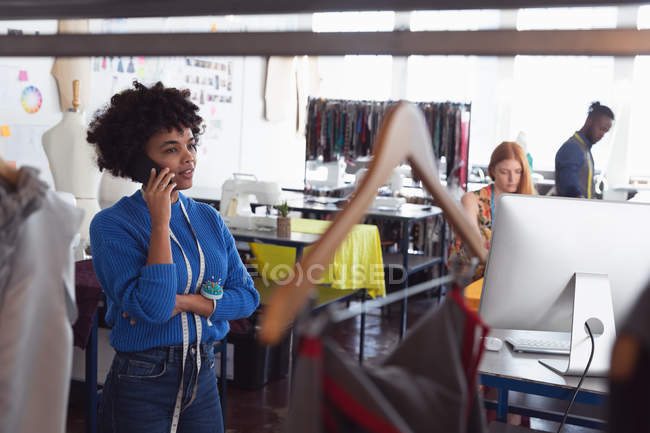 Vista frontal de una joven estudiante de moda afroamericana hablando en un teléfono inteligente en un estudio de la universidad de moda, con otros estudiantes trabajando en segundo plano - foto de stock