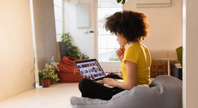 Vista laterale di una giovane donna di razza mista seduta su un sacchetto di fagioli con le gambe incrociate utilizzando un computer portatile in un ufficio creativo — Foto stock