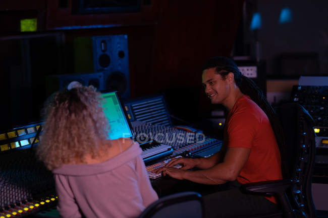 Vista lateral de cerca de un joven ingeniero de sonido masculino de raza mixta sentado y trabajando con una joven mujer caucásica con su espalda a la cámara en un escritorio de mezcla en un estudio de grabación - foto de stock