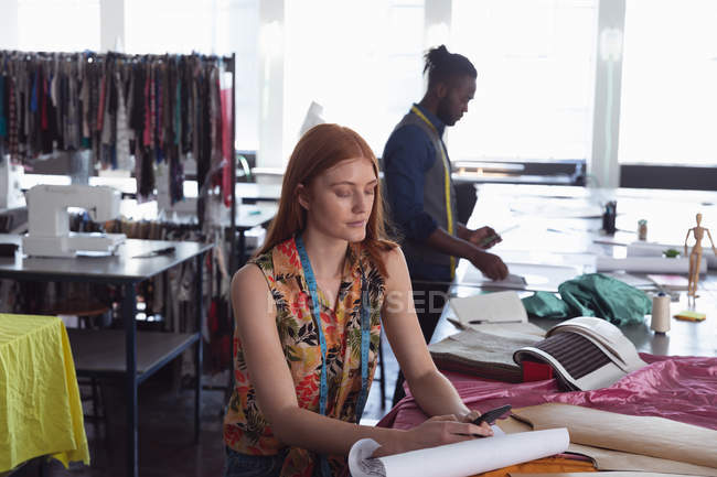 Vista frontal de una joven estudiante de moda caucásica trabajando en un diseño en un estudio en la universidad de moda, con un estudiante masculino trabajando en el fondo - foto de stock