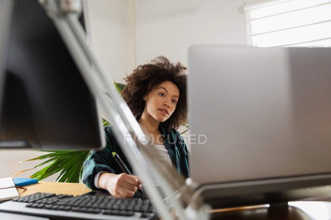 Вид спереду крупним планом молода змішана жінка, що сидить за столом, використовуючи ноутбук у творчому офісі — стокове фото