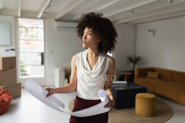 Вид спереди на молодую женщину смешанной расы, стоящую и держащую архитектурный рисунок в креативном офисе — стоковое фото