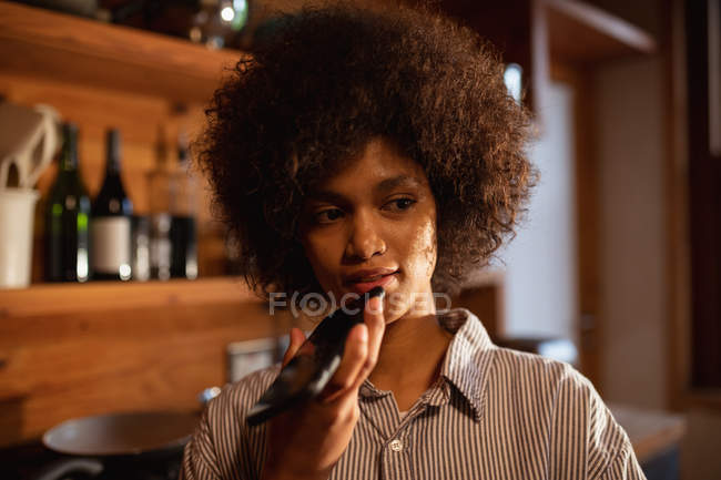 Вид спереду крупним планом молода змішана жінка в сорочці, що стоїть за допомогою смартфона вдома на її кухні — стокове фото