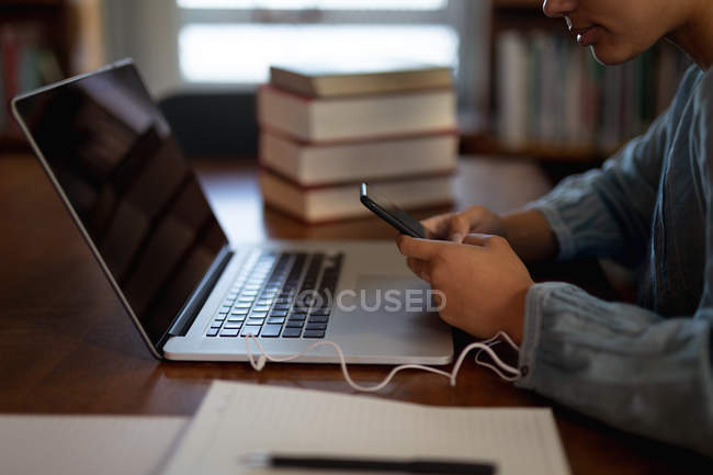 Vista lateral de perto de uma jovem estudante asiática usando um smartphone, um computador portátil e estudando em uma biblioteca — Fotografia de Stock