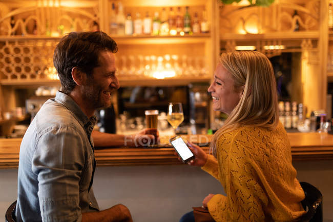 Vista lateral de um jovem casal caucasiano feliz relaxando juntos em férias em um bar, bebendo cerveja, vinho e usando um smartphone — Fotografia de Stock