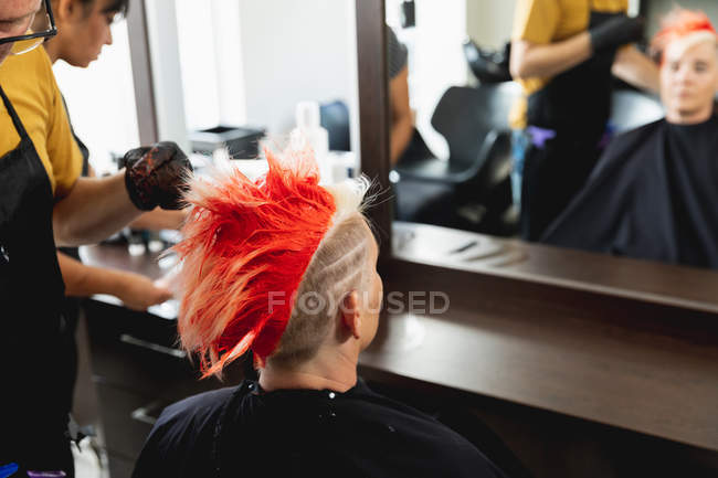 Передня картина середнього віку Кавказька перукарка і молода кавказька жінка з яскраво-червоним волоссям в салоні для волосся, відбитим у дзеркалі. — стокове фото