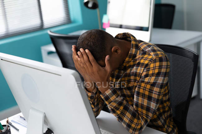 Vista frontal de un joven afroamericano sentado en un escritorio usando una computadora con la cabeza en las manos en la oficina moderna de un negocio creativo - foto de stock