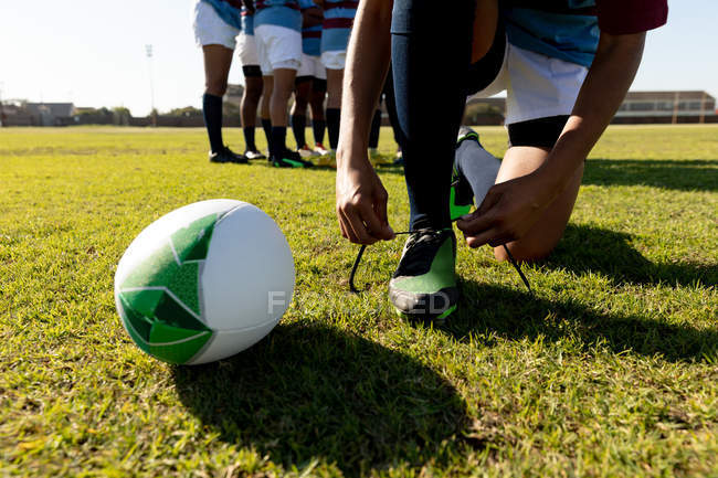 Front view low section of female rugby player kneeling and tying her boot on a rugby pitch, with her teammates talking together in the background — Stock Photo