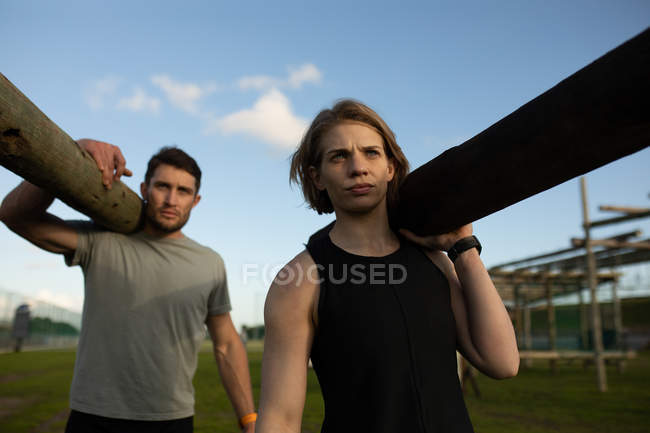 Front view of a young Caucasian woman and a young Caucasian man carrying logs of wood at an outdoor gym during a bootcamp training session — Stock Photo