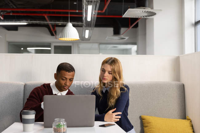 Vista frontal de perto de um jovem afro-americano e uma jovem mulher caucasiana sentada olhando para um computador portátil e conversando em uma mesa na área de jantar de um negócio criativo moderno — Fotografia de Stock
