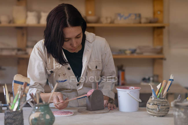 Front view close up of a young Caucasian female potter sitting at a work table painting a colored glaze on a clay flask in a pottery studio — Stock Photo