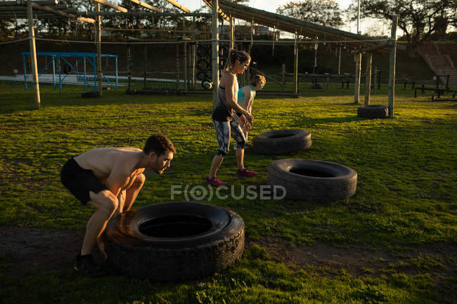 Vista lateral de dos jóvenes mujeres caucásicas y un joven hombre caucásico volteando neumáticos en un gimnasio al aire libre durante una sesión de entrenamiento de bootcamp - foto de stock