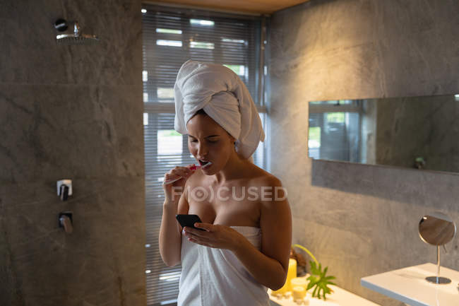 Вид спереди молодой белой женщины с завернутыми в полотенце волосами, чистящей зубы и смотрящей на свой смартфон в современной ванной комнате — стоковое фото